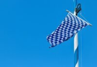 Blue-and-white checkered Bavarian flag fluttering from a mast against a clear blue sky