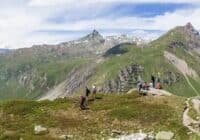 Panoramic mountain landscape with a trail and hikers along a ridge under a blue sky with distant snow-capped peaks.