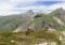 Panoramic mountain landscape with a trail and hikers along a ridge under a blue sky with distant snow-capped peaks.