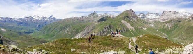 Panoramic mountain landscape with a trail and hikers along a ridge under a blue sky with distant snow-capped peaks.