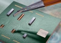 Close-up of a green printed circuit board with multiple small chips and gold contacts, as tweezers lift a connector from the board.