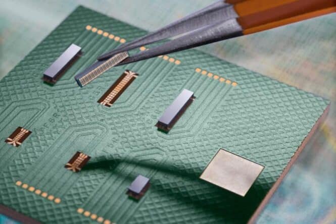 Close-up of a green printed circuit board with multiple small chips and gold contacts, as tweezers lift a connector from the board.