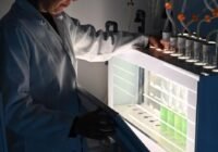 Researcher in a white lab coat loading sample tubes into a brightly lit laboratory analyzer.