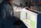 Researcher in a white lab coat loading sample tubes into a brightly lit laboratory analyzer.