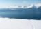 Snowy hillside with a single trail leading toward a blue sea and distant snow-covered mountains; a line of hikers follow the path in the foreground.