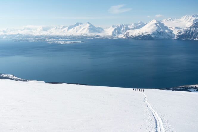 Snowy hillside with a single trail leading toward a blue sea and distant snow-covered mountains; a line of hikers follow the path in the foreground.