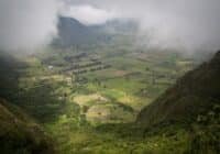 A misty valley with patchwork fields between green hills and distant mountains, partly obscured by low clouds.