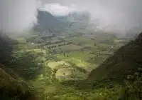 A misty valley with patchwork fields between green hills and distant mountains, partly obscured by low clouds.