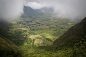 A misty valley with patchwork fields between green hills and distant mountains, partly obscured by low clouds.