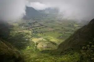 A misty valley with patchwork fields between green hills and distant mountains, partly obscured by low clouds.