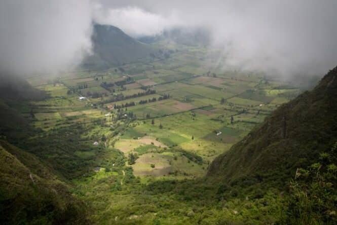 A misty valley with patchwork fields between green hills and distant mountains, partly obscured by low clouds.
