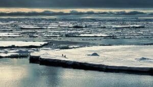 Frozen sea with large ice floes under a cloudy sky; two people walking on the ice edge.