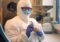 Scientist in full PPE (coveralls, mask, goggles) holding a brown paper bag with a sample in a laboratory setting.