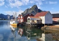 Harbor scene with a white and yellow fishing boat docked beside red wooden houses on stilts, mountains in the background.