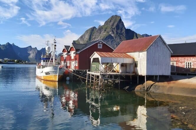 Harbor scene with a white and yellow fishing boat docked beside red wooden houses on stilts, mountains in the background.