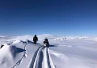 Person walking beside a snowmobile on a snow-covered, mountainous landscape under a clear blue sky.