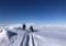 Person walking beside a snowmobile on a snow-covered, mountainous landscape under a clear blue sky.