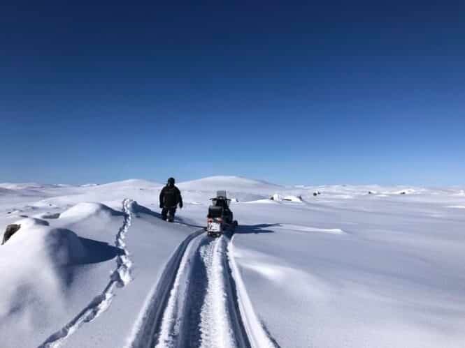 Person walking beside a snowmobile on a snow-covered, mountainous landscape under a clear blue sky.