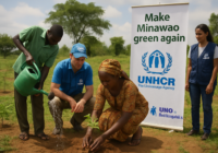 Group of volunteers planting a sapling beside a UNHCR banner that reads 'Make Minawao green again' in a rural field.