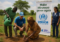 Group of volunteers planting a sapling beside a UNHCR banner that reads 'Make Minawao green again' in a rural field.