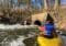 Kayaker in a yellow boat wearing a blue life jacket paddles toward a concrete culvert in a fast-flowing river with trees along the bank.