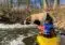 Kayaker in a yellow boat wearing a blue life jacket paddles toward a concrete culvert in a fast-flowing river with trees along the bank.