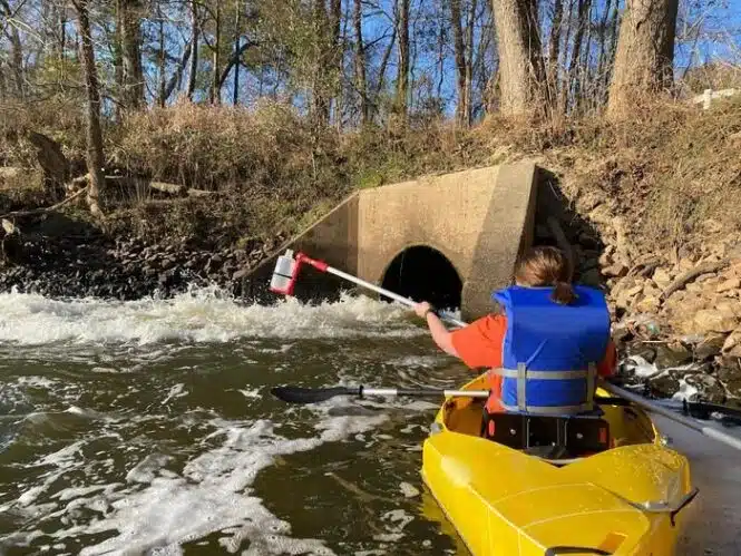 Kayaker in a yellow boat wearing a blue life jacket paddles toward a concrete culvert in a fast-flowing river with trees along the bank.