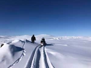 Person in winter gear walking beside a snowmobile along a snow-covered plain under a clear blue sky.