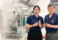 Two Illinois researchers in navy polo shirts holding small vials beside a large lab instrument, smiling at the camera.