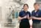 Two Illinois researchers in navy polo shirts holding small vials beside a large lab instrument, smiling at the camera.