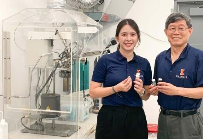 Two Illinois researchers in navy polo shirts holding small vials beside a large lab instrument, smiling at the camera.