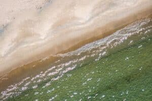 Aerial view of a sandy beach meeting clear green water with gentle waves and sunlight glints.