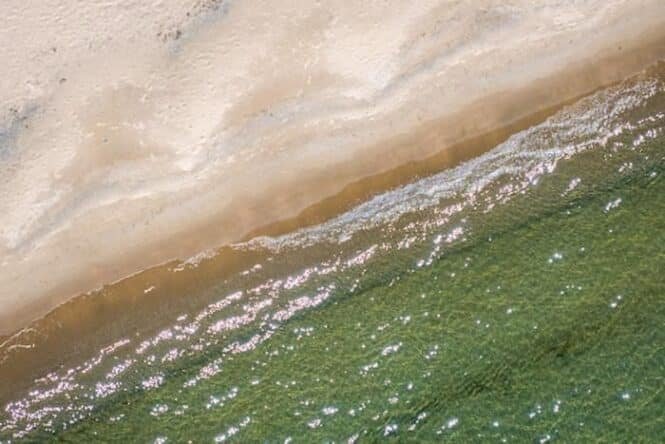 Aerial view of a sandy beach meeting clear green water with gentle waves and sunlight glints.