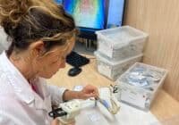Dental lab technician in a white coat works on a dental model with a handheld tool at a workstation, computer and labeled plastic bins in the background.