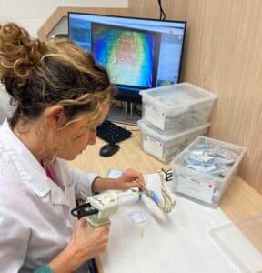 Dental lab technician in a white coat works on a dental model with a handheld tool at a workstation, computer and labeled plastic bins in the background.
