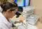 Dental lab technician in a white coat works on a dental model with a handheld tool at a workstation, computer and labeled plastic bins in the background.