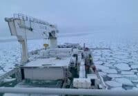 Ice-covered sea viewed from a cargo ship deck, with a large gantry crane on the left and frozen ice floes stretching to the horizon; crew members in orange jackets are visible on deck.