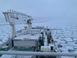 Ice-covered sea viewed from a cargo ship deck, with a large gantry crane on the left and frozen ice floes stretching to the horizon; crew members in orange jackets are visible on deck.
