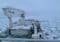Ice-covered sea viewed from a cargo ship deck, with a large gantry crane on the left and frozen ice floes stretching to the horizon; crew members in orange jackets are visible on deck.