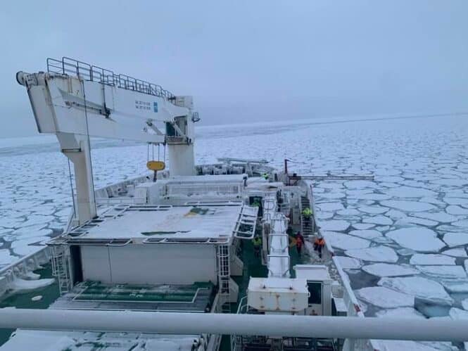 Ice-covered sea viewed from a cargo ship deck, with a large gantry crane on the left and frozen ice floes stretching to the horizon; crew members in orange jackets are visible on deck.