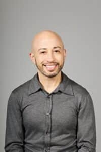 Portrait of a smiling bald man wearing a gray button-up shirt, posed against a neutral gray background.