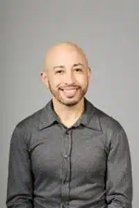 Portrait of a smiling bald man wearing a gray button-up shirt, posed against a neutral gray background.