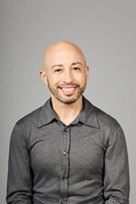 Portrait of a smiling bald man wearing a gray button-up shirt, posed against a neutral gray background.