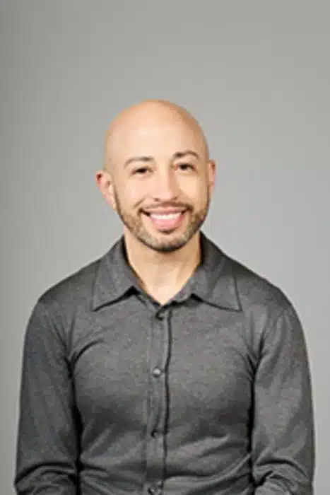 Portrait of a smiling bald man wearing a gray button-up shirt, posed against a neutral gray background.