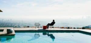 Person relaxing on a lounge chair beside a rooftop pool, overlooking a distant city skyline.