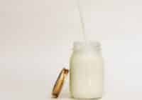 Milk is being poured into a glass mason jar with a metal lid resting on its side on a pale background.