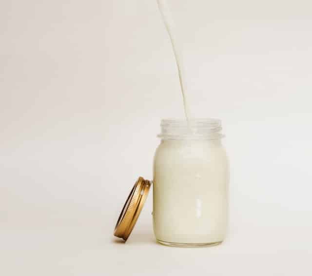 Milk is being poured into a glass mason jar with a metal lid resting on its side on a pale background.