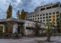Abandoned hotel with cracked concrete facade, overgrown trees, and a statue near the entrance.