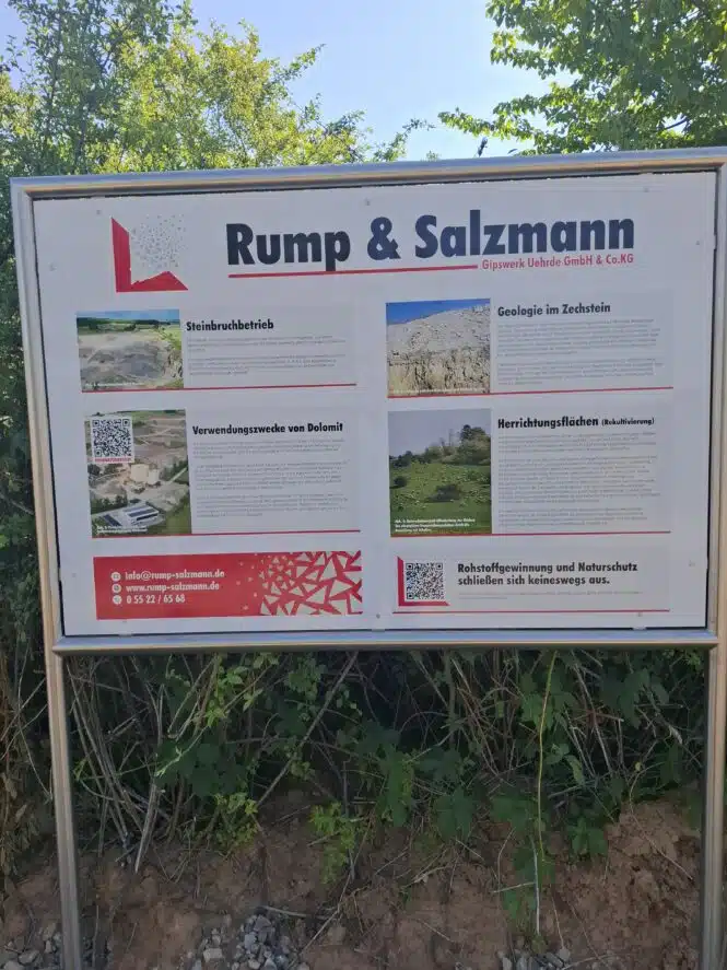 Outdoor information sign for Rump & Salzmann (Gipswerk Uehrde GmbH & Co. KG) with sections on stone quarrying and geology, images and descriptive text in German, mounted in a metal frame beside the path.