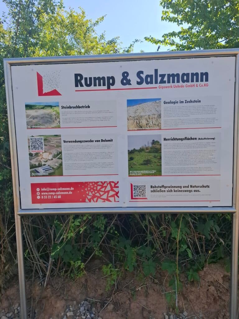Outdoor information sign for Rump & Salzmann (Gipswerk Uehrde GmbH & Co. KG) with sections on stone quarrying and geology, images and descriptive text in German, mounted in a metal frame beside the path.